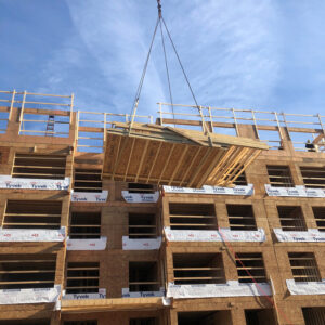 A crane lifts a large wooden frame panel to the upper floors of a multi-story building under construction, with blue sky and scattered clouds in the background.
