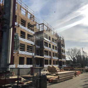 A multi-story building under construction, with exposed wooden framing and scaffolding. Construction materials are stacked on the ground, and workers can be seen on upper floors. The sky is partly cloudy with sunlight streaming in.