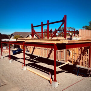 A building under construction with a red steel frame and wooden structures, set on a gravel lot. Some houses and trees are visible in the background under a clear blue sky.