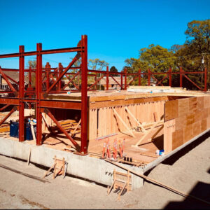 A building under construction with exposed red steel beams and wooden framing, set on a concrete foundation. Construction materials and tools are scattered around the site. Trees and houses are visible in the background under a clear blue sky.