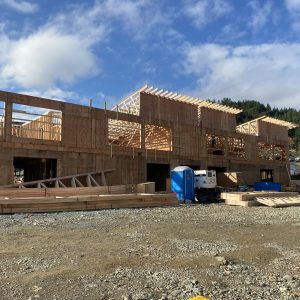 A multi-story wooden building frame under construction on a sunny day. Piles of lumber are in the foreground.