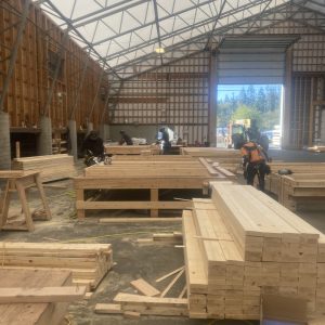 Several people work with stacks of lumber inside a large, open industrial warehouse with high ceilings and a partially open garage door. Construction tools and materials are scattered throughout the workspace.
