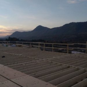 A building under construction with exposed wooden beams in the foreground, set against a backdrop of mountains and a sunset sky.