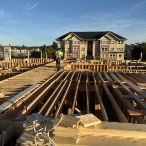 Construction workers on a building site frame a new structure with a finished apartment building in the background.