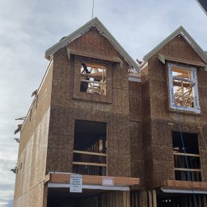 Two unfinished three-story houses with exposed wooden framing, missing windows, and a crane lifting construction materials to the roof. Signs and building materials are visible at ground level. The sky is partly cloudy.