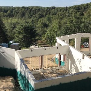 A partially constructed house foundation with white and green walls stands on a dirt lot surrounded by dense, green forest and distant hills under a clear sky. Several ladders lean against the structure.