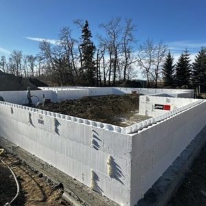 A construction site shows the foundation of a building made from white insulated concrete forms, surrounded by dirt piles and trees under a clear blue sky.