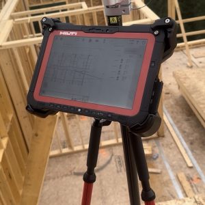 A Hilti tablet on a tripod displays construction plans at a building site with wooden framing in progress and trees in the background.