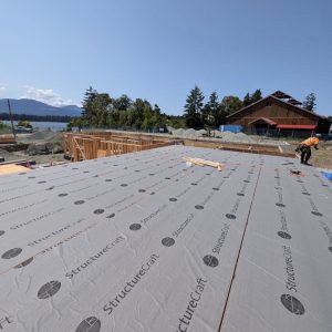 Construction site with a partially built wooden structure, workers in safety gear, and a large surface covered with StructureCraft panels. Trees, buildings, and a mountain range are visible in the background under a clear sky.