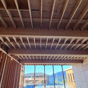 View from inside a building under construction, showing exposed wooden beams, framing, and an open ceiling. Through the opening, other buildings and a mountain are visible in the background.