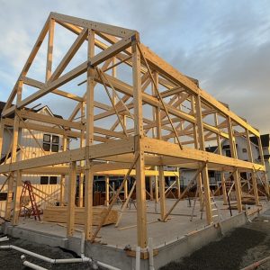 A wooden framework of a house under construction is seen on a concrete foundation at sunset, with residential buildings and construction materials in the background.