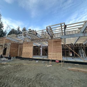 Partially constructed building with exposed wooden beams, plywood walls, and open framework. Construction materials and equipment are scattered on the ground, with trees and a blue sky in the background.
