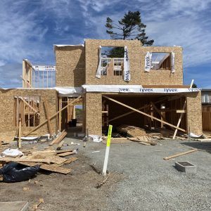 A two-story house under construction, framed with wooden beams and OSB sheathing. Building materials, debris, and tools are scattered around the gravel lot. Some Tyvek house wrap is visible on the upper level.