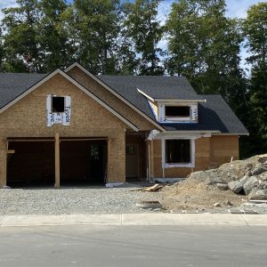 A house under construction with unfinished exterior walls, garage, and exposed windows, surrounded by dirt, rocks, and construction materials, with trees in the background.