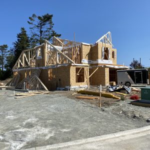 A two-story house under construction with wooden framing and exposed plywood, surrounded by building materials and equipment on a clear, sunny day.