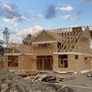 A house under construction with wooden framing and exposed beams, set against a partly cloudy sky. Construction materials and dirt piles are visible in the foreground.