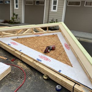 A large triangular roof truss lies on the ground at a construction site, with tools and materials nearby. The truss is covered with building wrap labeled "Dupont" and is in front of modern garages.