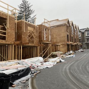 Row of townhouses under construction on a snowy day, with wooden framing exposed and building materials scattered along the roadside. Some houses are further along in construction, while others are still being framed.