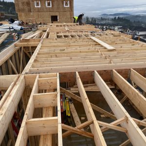 Wooden framing of a house under construction, with workers on the site and a partially built structure in the background. Misty hills and neighboring houses are visible in the distance.