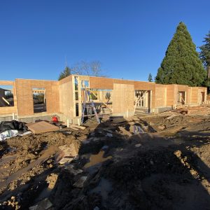 A construction site with the wooden frame of a single-story building under clear blue skies, surrounded by mud, scattered materials, and tall trees in the background.