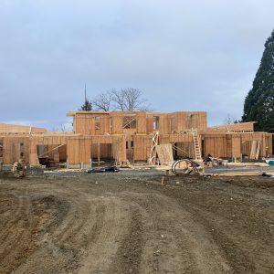 A two-story wooden house is under construction on a dirt lot, with framing partially completed and construction materials scattered around. A large evergreen tree is visible to the right.