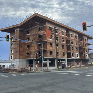 A multi-story building under construction at a street corner, with wooden framing visible and scaffolding around the structure. Traffic lights and road signs are present in the foreground under a cloudy sky.