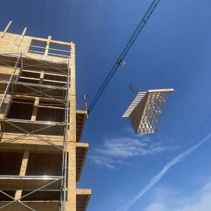 A construction crane lifts a large wooden framework panel beside a multi-story building under construction, with scaffolding and a clear blue sky in the background.