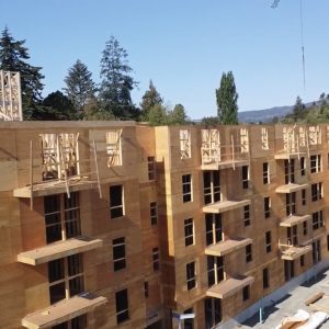 A multi-story wooden apartment building under construction, with open window frames, exposed wood beams, balconies, and construction materials and cranes visible around the site. Trees and blue sky are in the background.