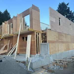 A two-story house under construction, with wooden framing and plywood walls partially completed, sits on a concrete foundation. Building materials and tools are scattered around the site. Trees and a neighboring building are in the background.