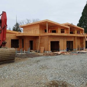A two-story house under construction with wooden framing, surrounded by construction materials and debris. An excavator is parked on the left, and gravel covers the ground in front of the building. Trees are visible in the background.