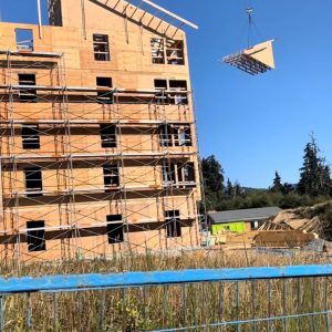A crane lifts a triangular wooden roof section toward the top of a partially constructed multi-story wooden building, surrounded by scaffolding and grassy field under a clear blue sky.