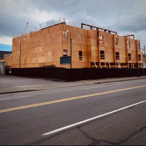 A large multi-story building under construction, framed and covered with plywood, stands on a corner lot near empty streets. The sky is cloudy and there is fencing around the construction site.