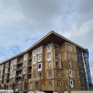 A multi-story apartment building under construction, with exposed wooden framing, unfinished windows, and scaffolding on one side, set against a cloudy sky.