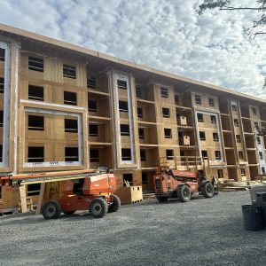 A large, multi-story building under construction with exposed wooden framing, multiple rectangular window openings, and construction equipment parked in front on a gravel-covered area under a partly cloudy sky.