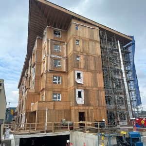 A multistory wooden building under construction, with scaffolding on one side and exposed plywood walls. Construction equipment, cones, and barriers are visible in the foreground under a cloudy sky.