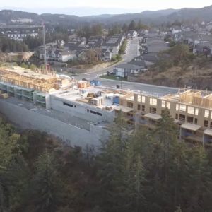 Aerial view of a large building under construction near a forested area, with wooden framing visible and a residential neighborhood in the background.