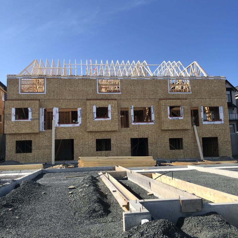 Construction of a multi-unit building with wood framing, OSB sheathing, and exposed roof trusses. The foreground shows gravel and concrete foundation work.