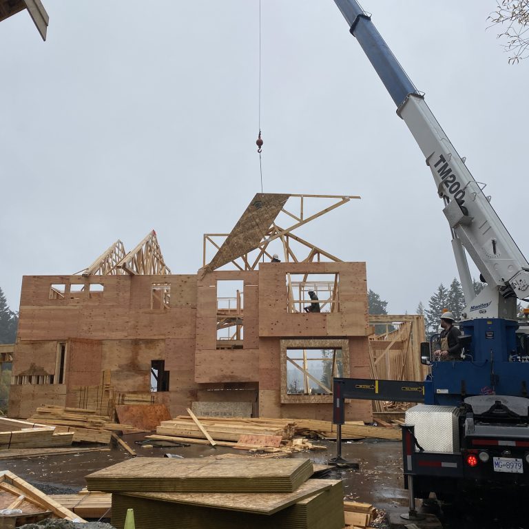 A crane lifts a wooden roof panel onto a partially constructed two-story house on a cloudy day. Construction materials and workers are visible around the site.