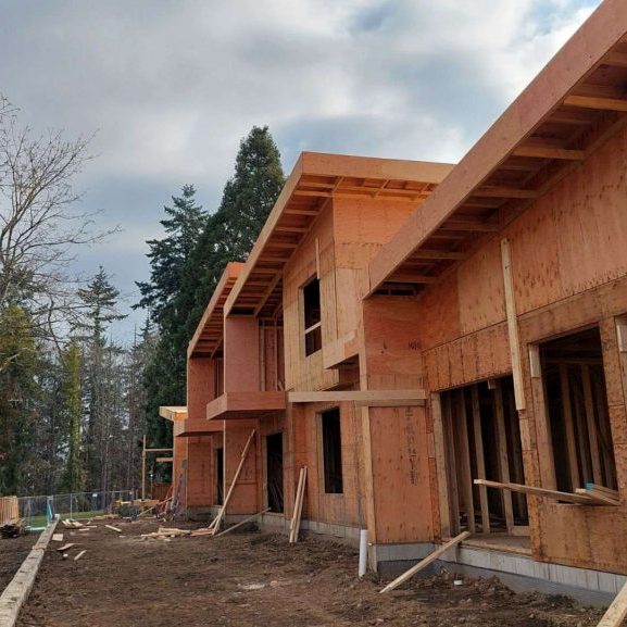 Partially constructed wooden house with exposed framing, large window openings, and a sloped roof. The site is surrounded by trees, dirt, and construction materials under a cloudy sky.